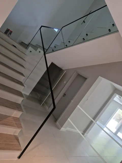 Glass along the staircase: Clear glass panels follow the staircase and landing, paired with a dark handrail and visible point fixings.
